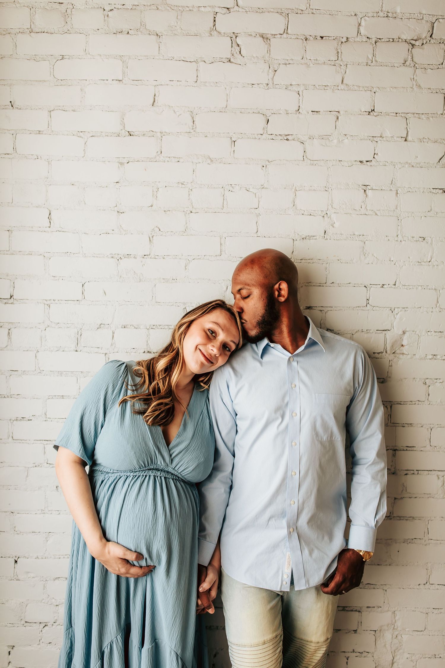 Expectant couple in soft blue outfits shares an intimate moment against a white brick wall.