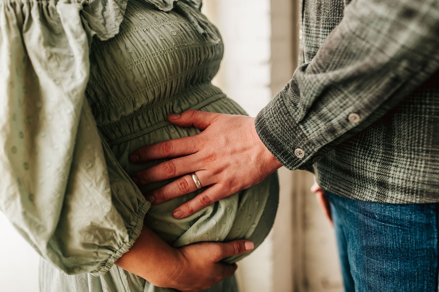 Close up of hands cradling a pregnant belly in olive green dress, with denim visible to the side.