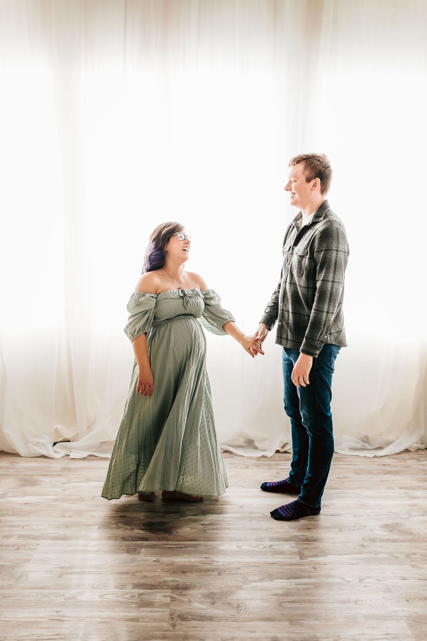 A couple holds hands in a light-filled studio, with the woman wearing a flowing sage green dress against a white backdrop.