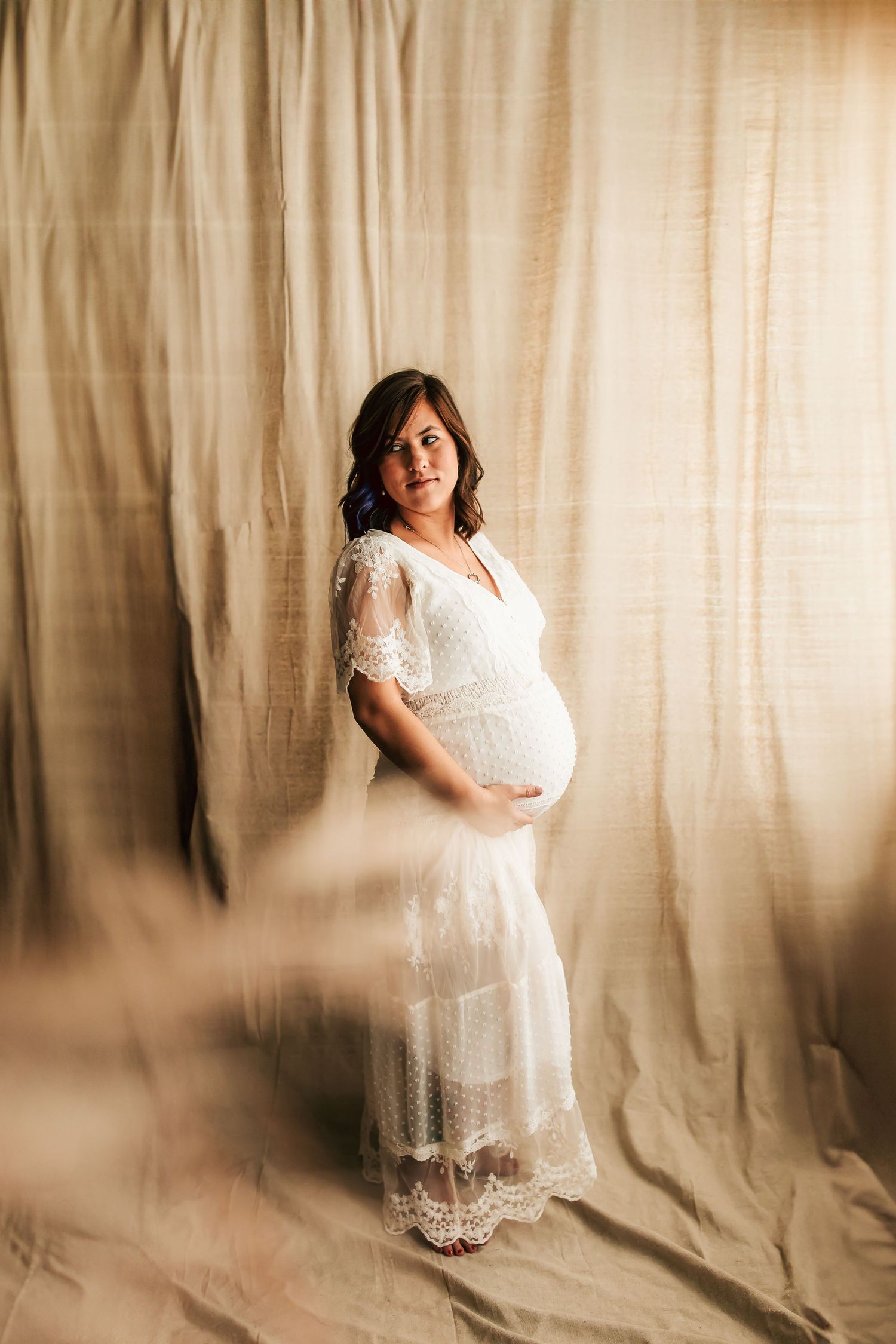 A pregnant woman poses in a white lace maternity dress against a beige backdrop for a studio portrait.