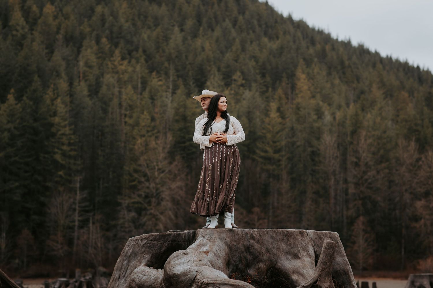 Woman in a long plaid skirt and white top stands on a large tree stump overlooking a forested mountain.