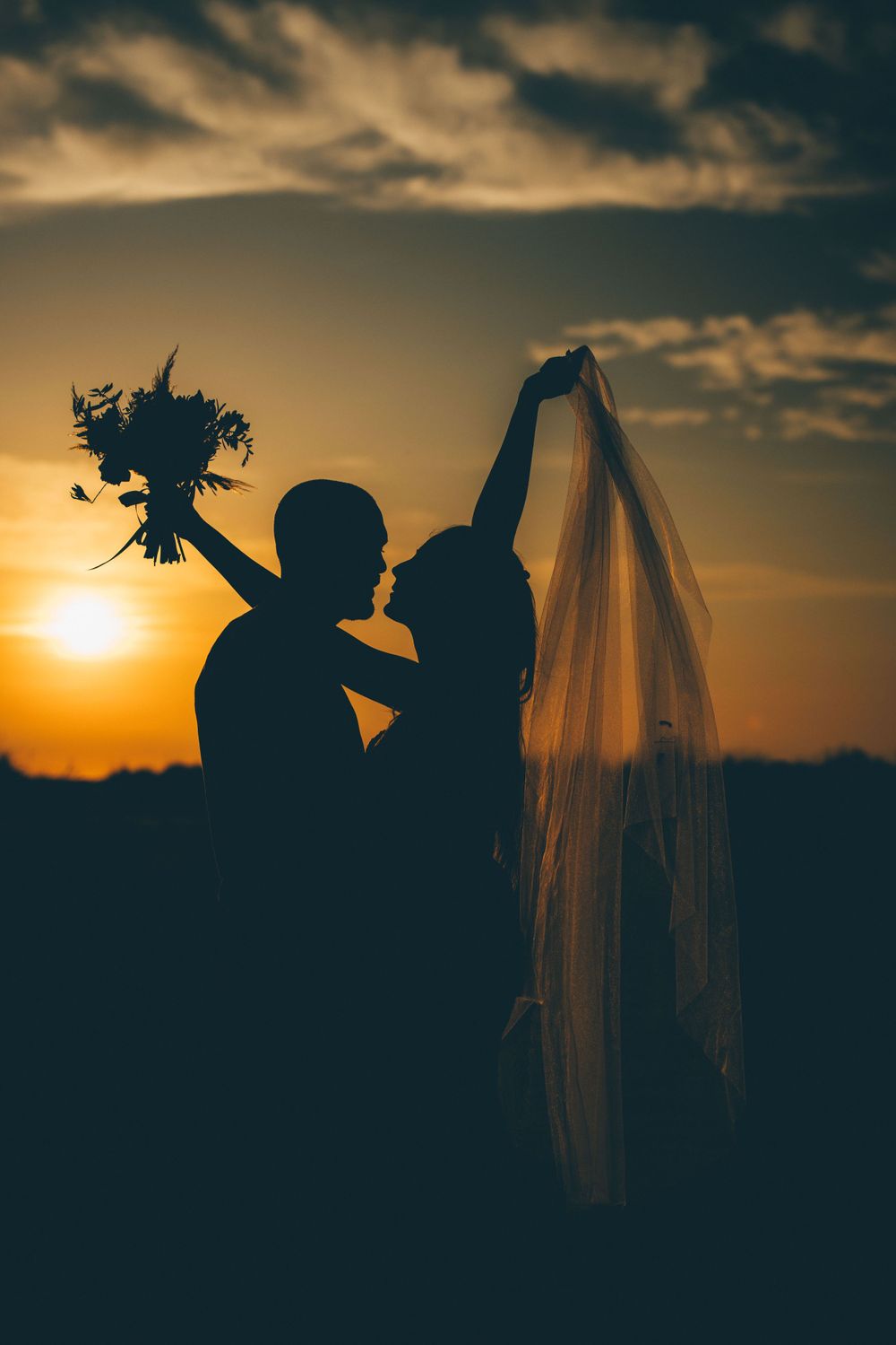 Silhouette de jeunes mariés tenant un bouquet et un voile sur fond de coucher de soleil spectaculaire.