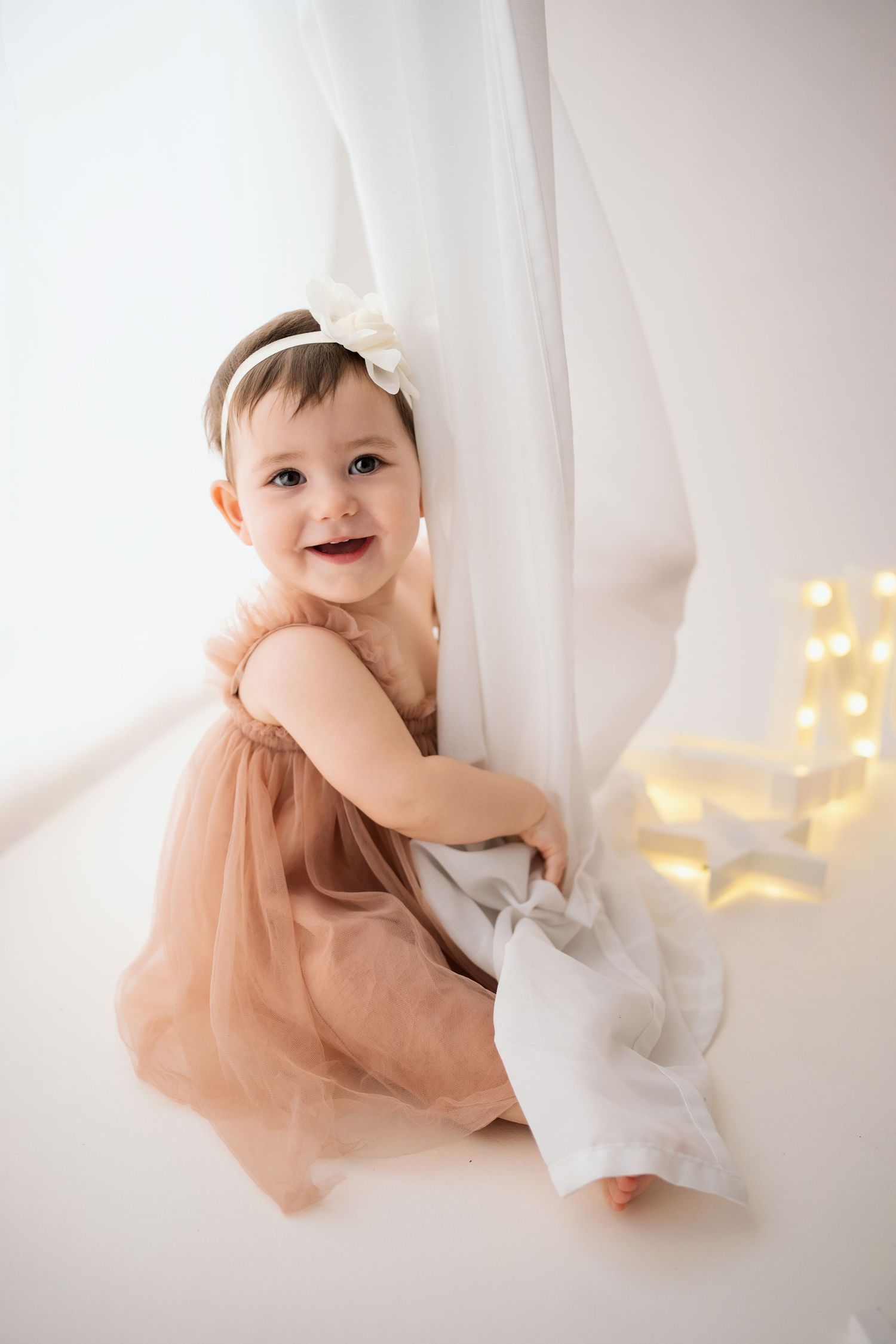 Happy baby girl in tutu dress with bow headband sitting by white curtain with fairy lights.