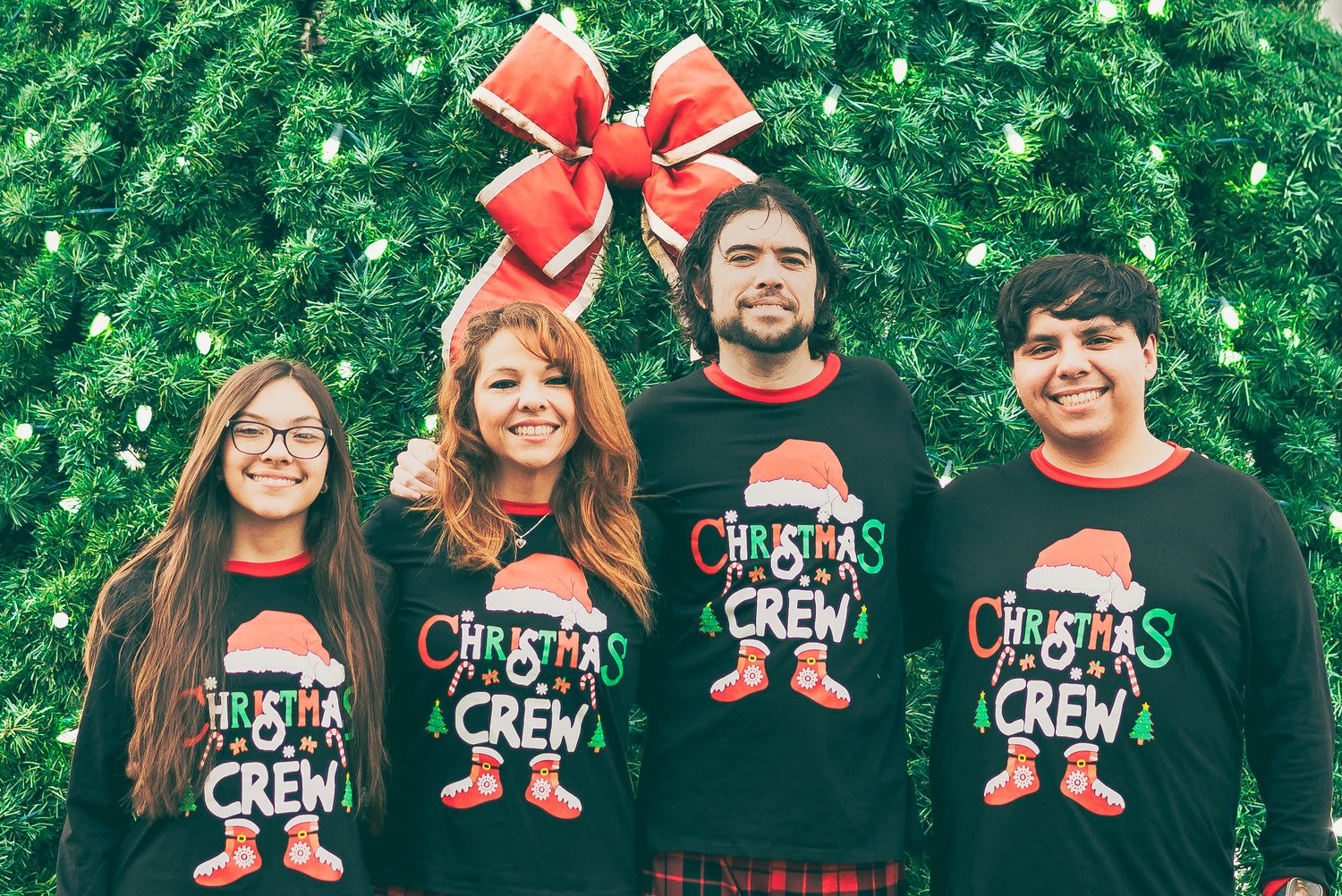 A group wearing matching Christmas crew graphic shirts with a red bow and green Christmas tree in the background.