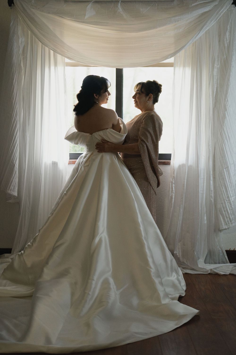 Two figures embrace in a sunlit room with flowing white wedding dress and sheer curtains creating a romantic atmosphere.