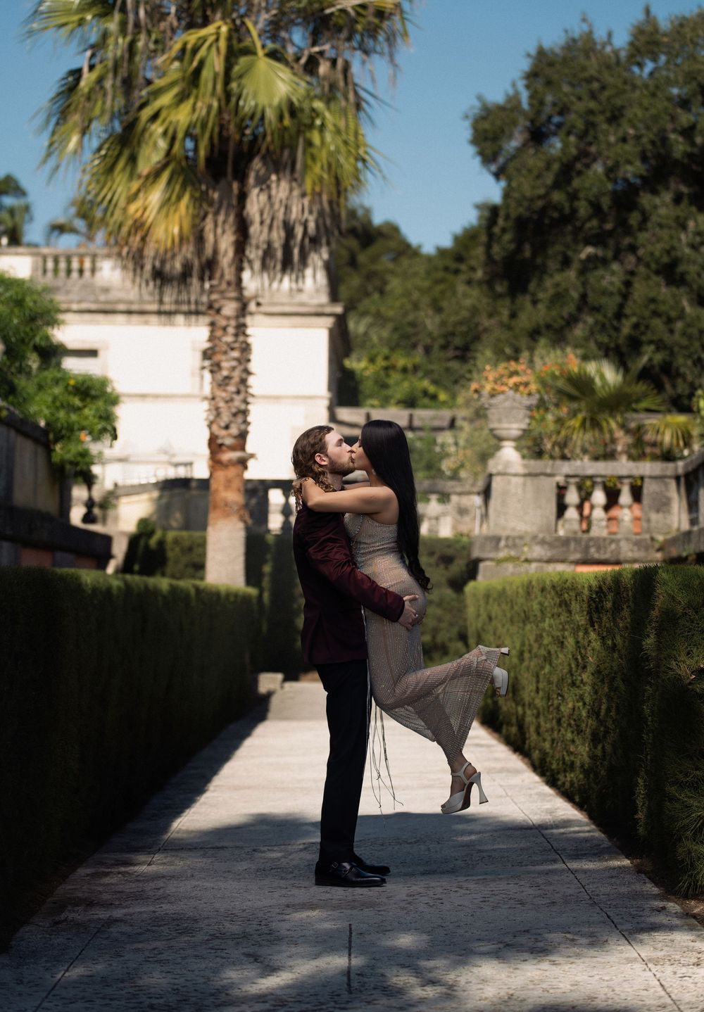 A romantic couple embraces in a garden pathway lined with palm trees and classical architecture.