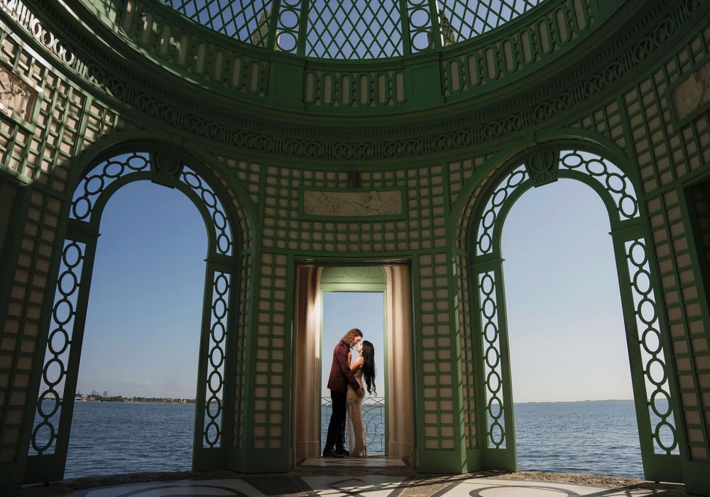Romantic silhouette in an ornate gazebo overlooking the ocean, framed by two arched windows with decorative lattice patterns.