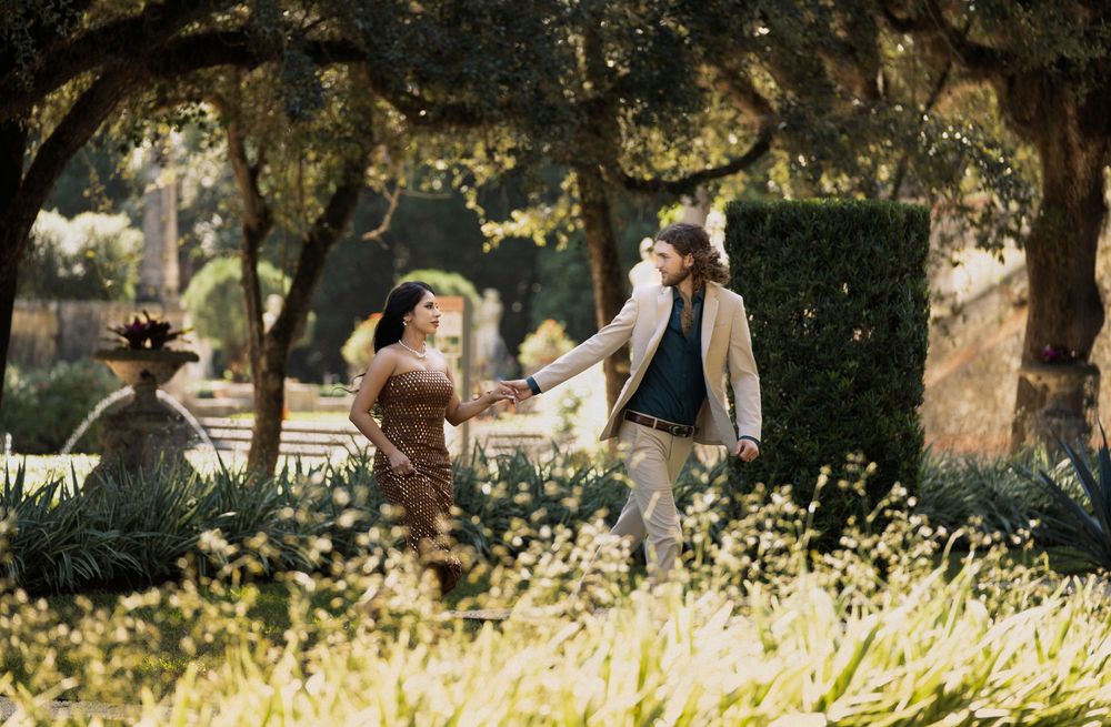 A couple walks together through a sunlit park pathway surrounded by lush trees and greenery.