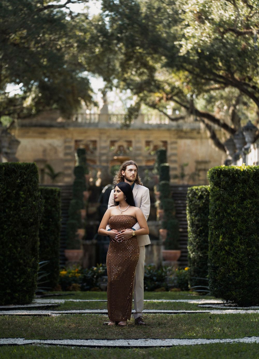 A couple embraces in a formal garden setting with tall hedges and classical architecture in the background.