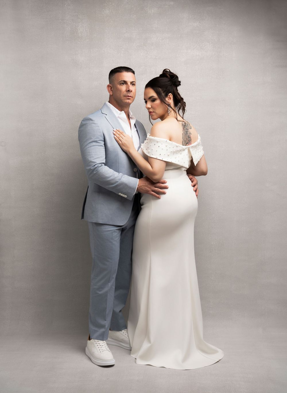 A couple poses together for a wedding photo wearing a light gray suit and off-shoulder white gown against a neutral backdrop.