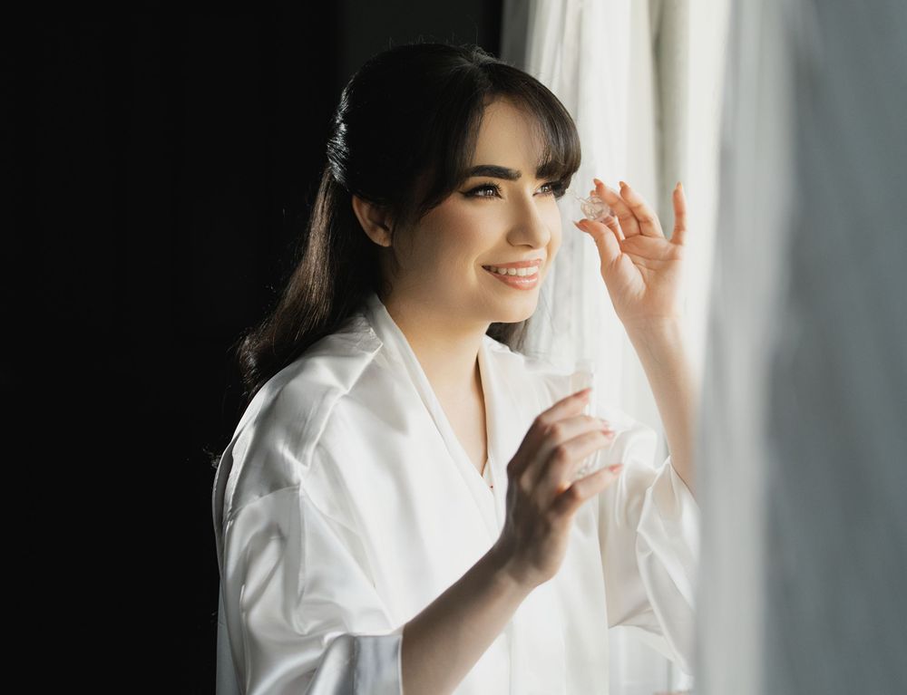 A person in a white robe sits by a window during morning preparations with soft natural light filtering in.