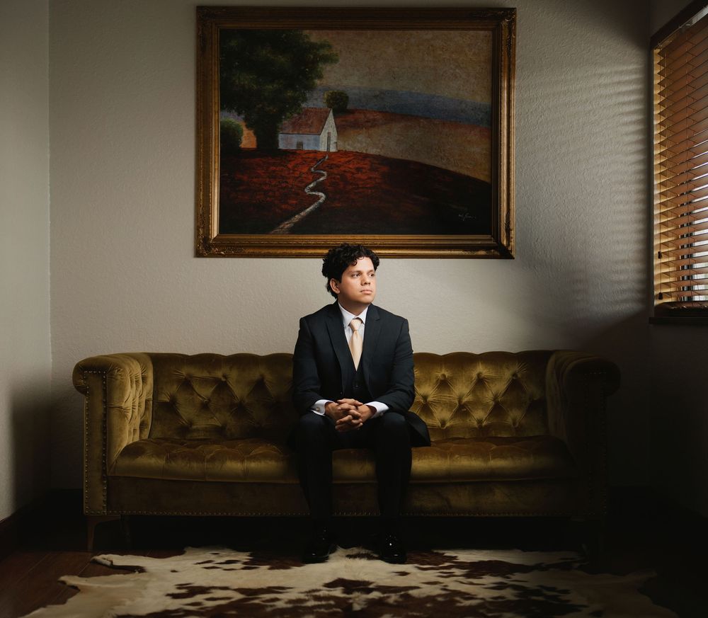 A person in a dark suit sits on a vintage gold couch beneath a framed landscape painting in moody lighting.