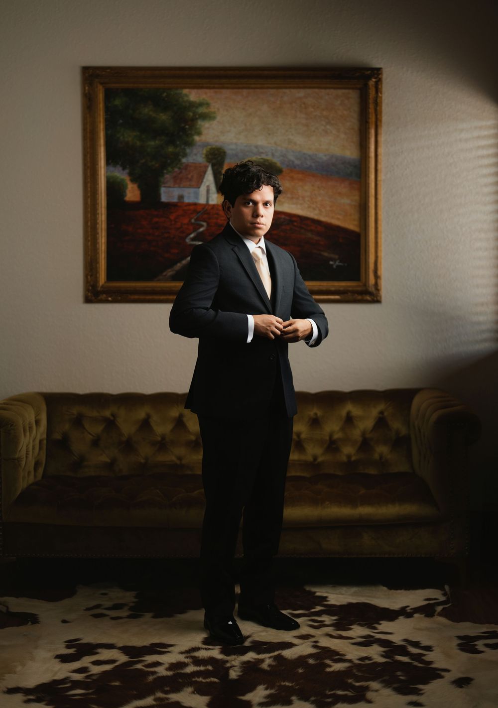 A formal portrait in a room with a vintage couch, landscape painting, and cowhide rug in moody lighting.