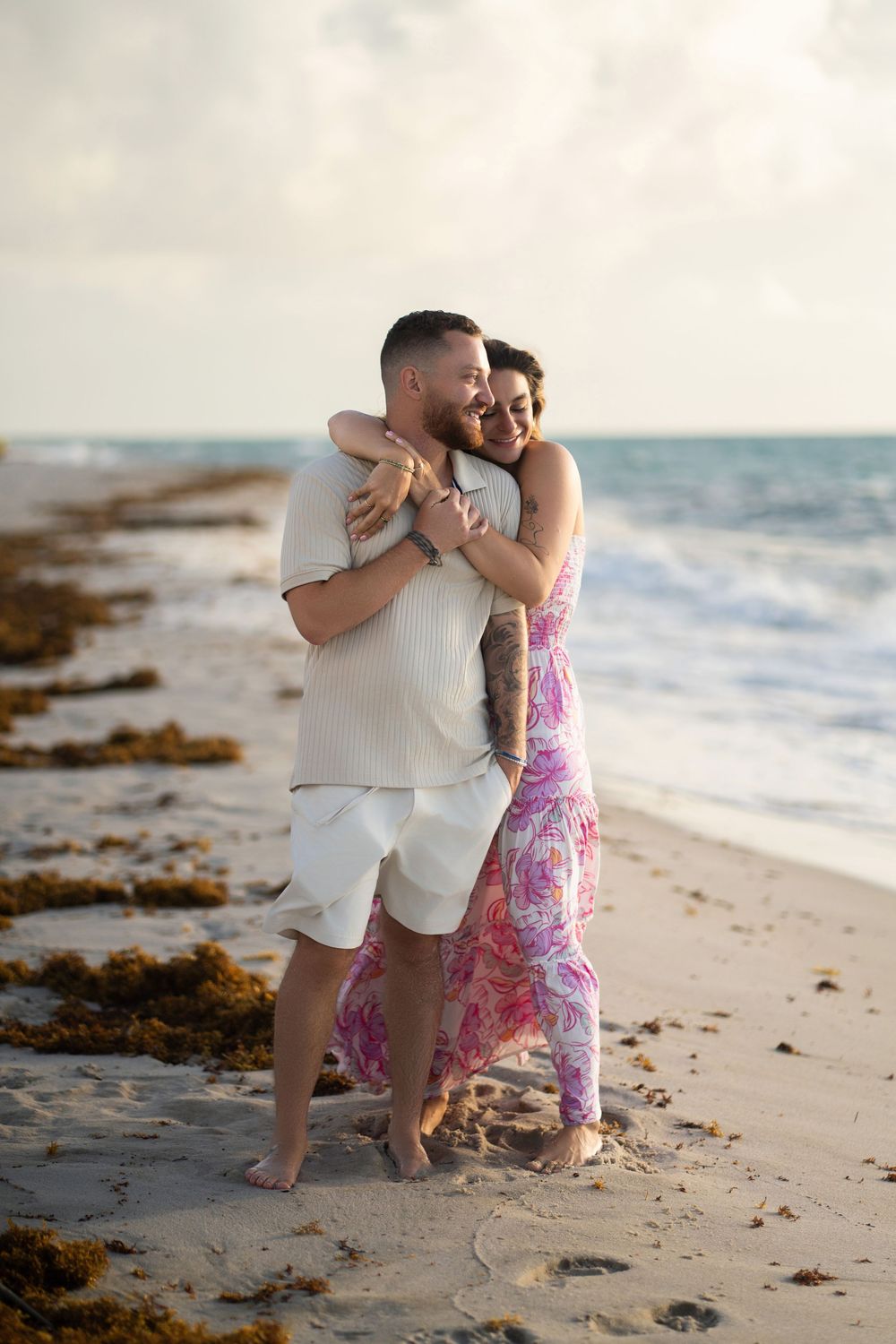 A couple embraces lovingly on a sandy beach at sunset while ocean waves crash in the background.