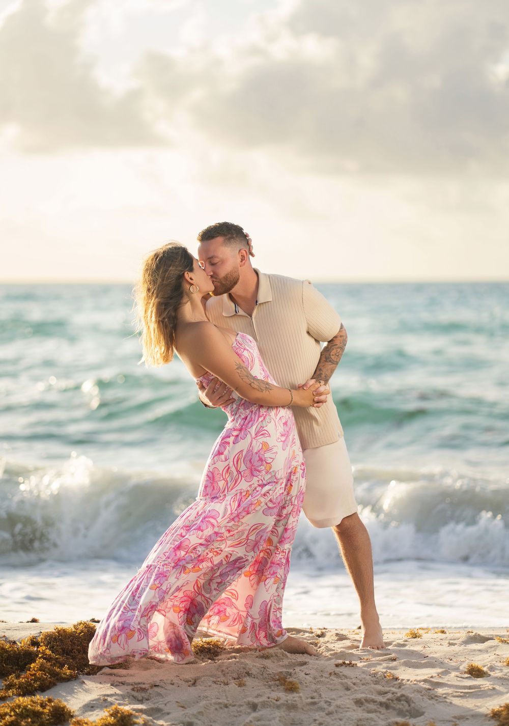 A couple in love embraces romantically on a sandy beach at sunset with ocean waves in the background.