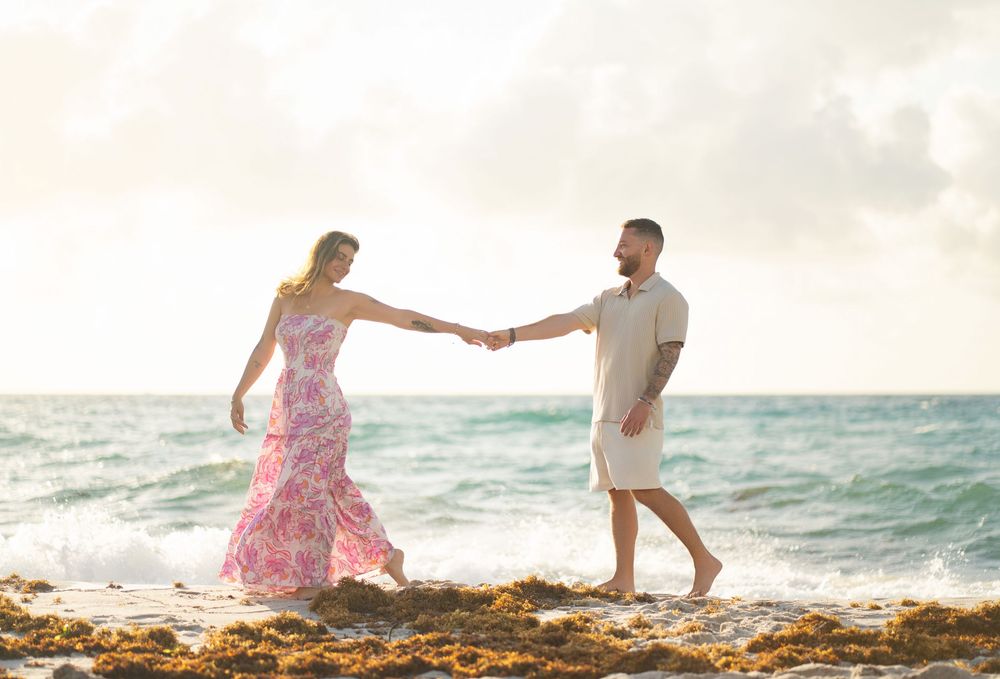 A couple holds hands on rocky coastal shore with turquoise waves and sunset sky in background.