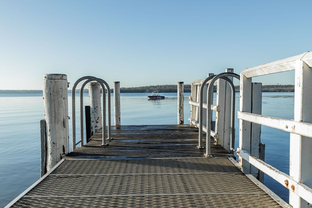 Wooden pier with white railings extends into calm blue waters with a small boat visible in the distance. Jervis Bay NSW