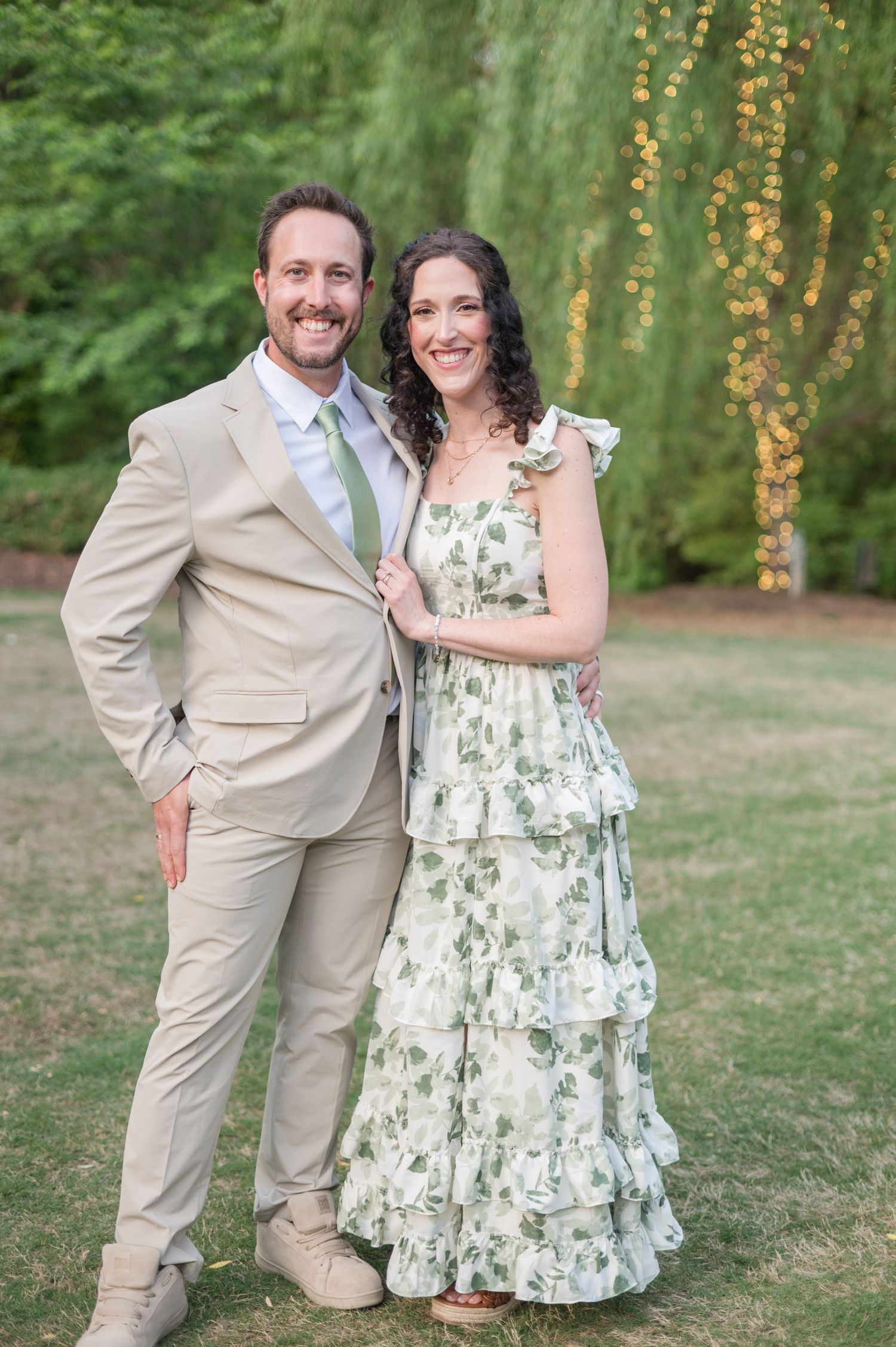 A couple embraces by a lake wearing summer attire - a coral off-shoulder top and white shirt - on a sunny afternoon.