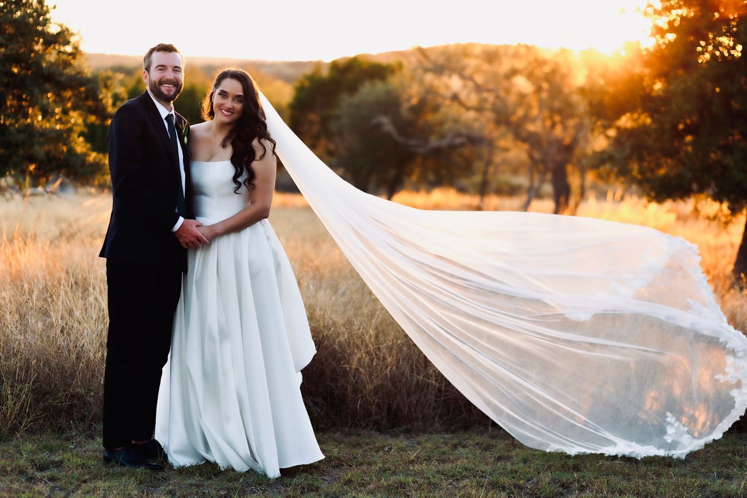Bride and groom at sunset with flowing veil in field.