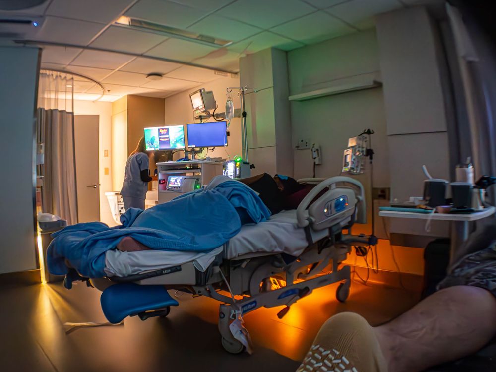 A hospital room with dim lighting shows a patient resting on a medical bed with monitors above.