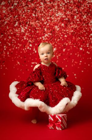 Small child in red tulle dress sits among christmas gifts and glittery decorations against festive red background.