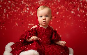 Baby in sparkly red dress sits against festive red background with falling snow effect.