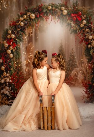 Two girls in champagne ball gowns pose by a festive Christmas arch with gold ornaments and red berries on a sled.