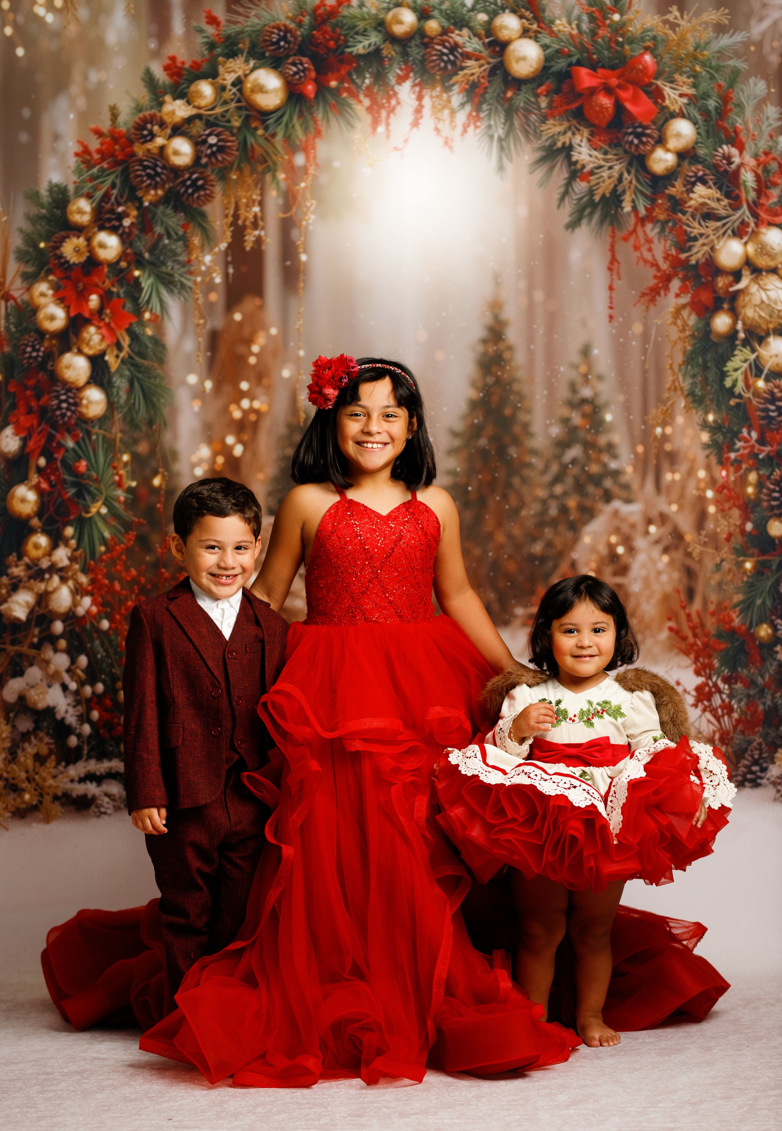 Three children in red holiday outfits pose under a festive Christmas wreath arch with ornaments and greenery.