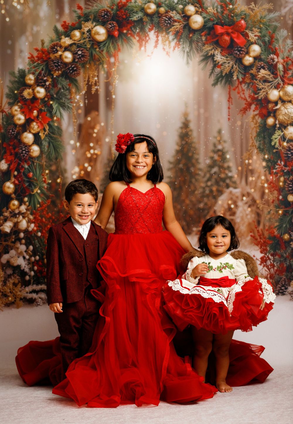 Three children in red holiday outfits pose under a festive Christmas wreath arch with ornaments and greenery.