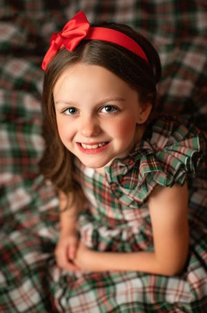 Holiday portrait with plaid dress and red bow headband against festive background.