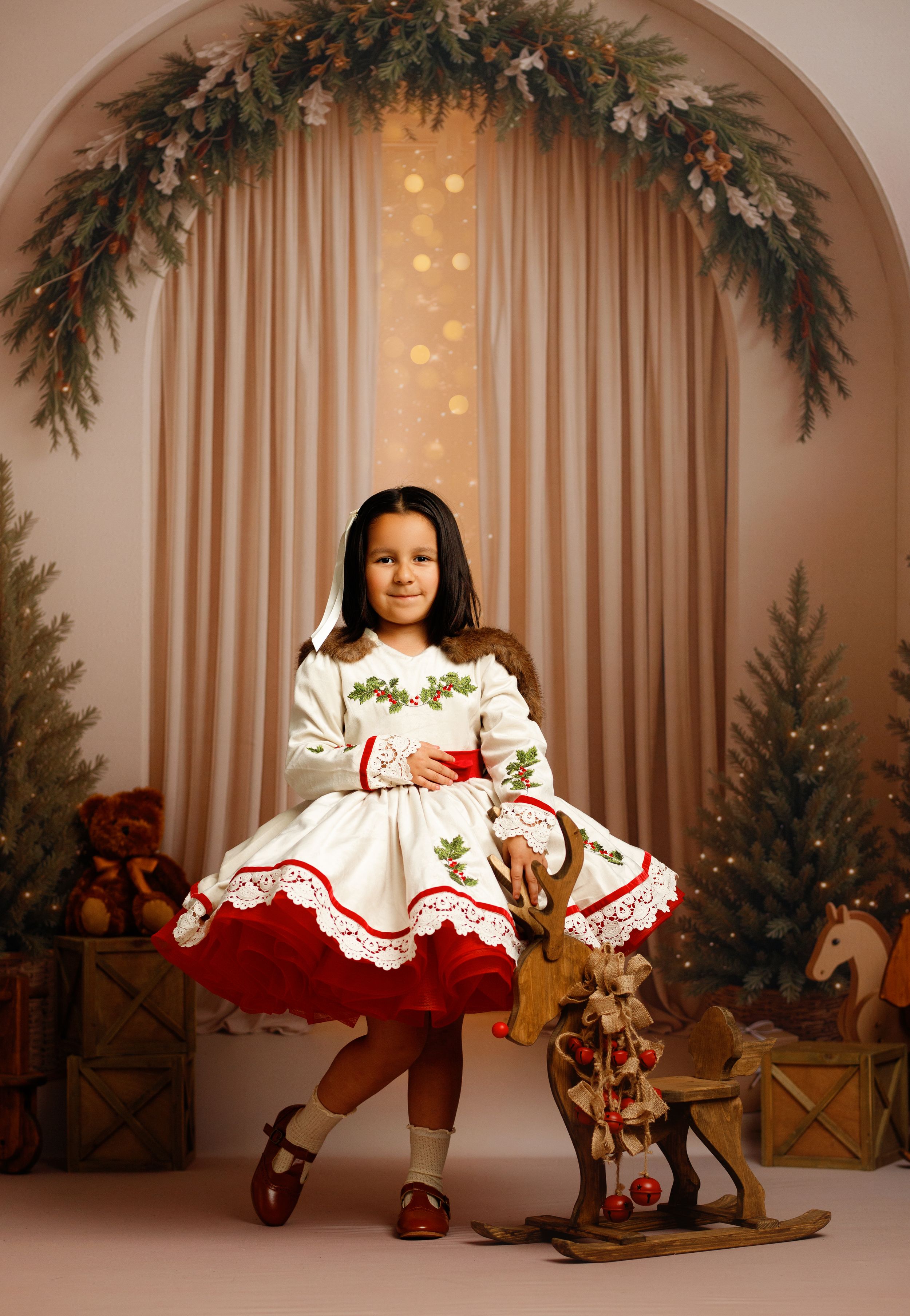 Young girl in white and red Christmas dress poses with wooden rocking horse in festive holiday portrait setting.
