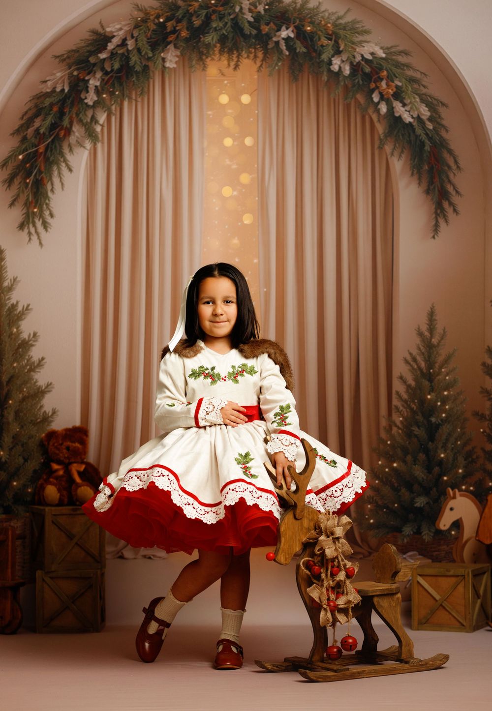 Young girl in white and red Christmas dress poses with wooden rocking horse in festive holiday portrait setting.