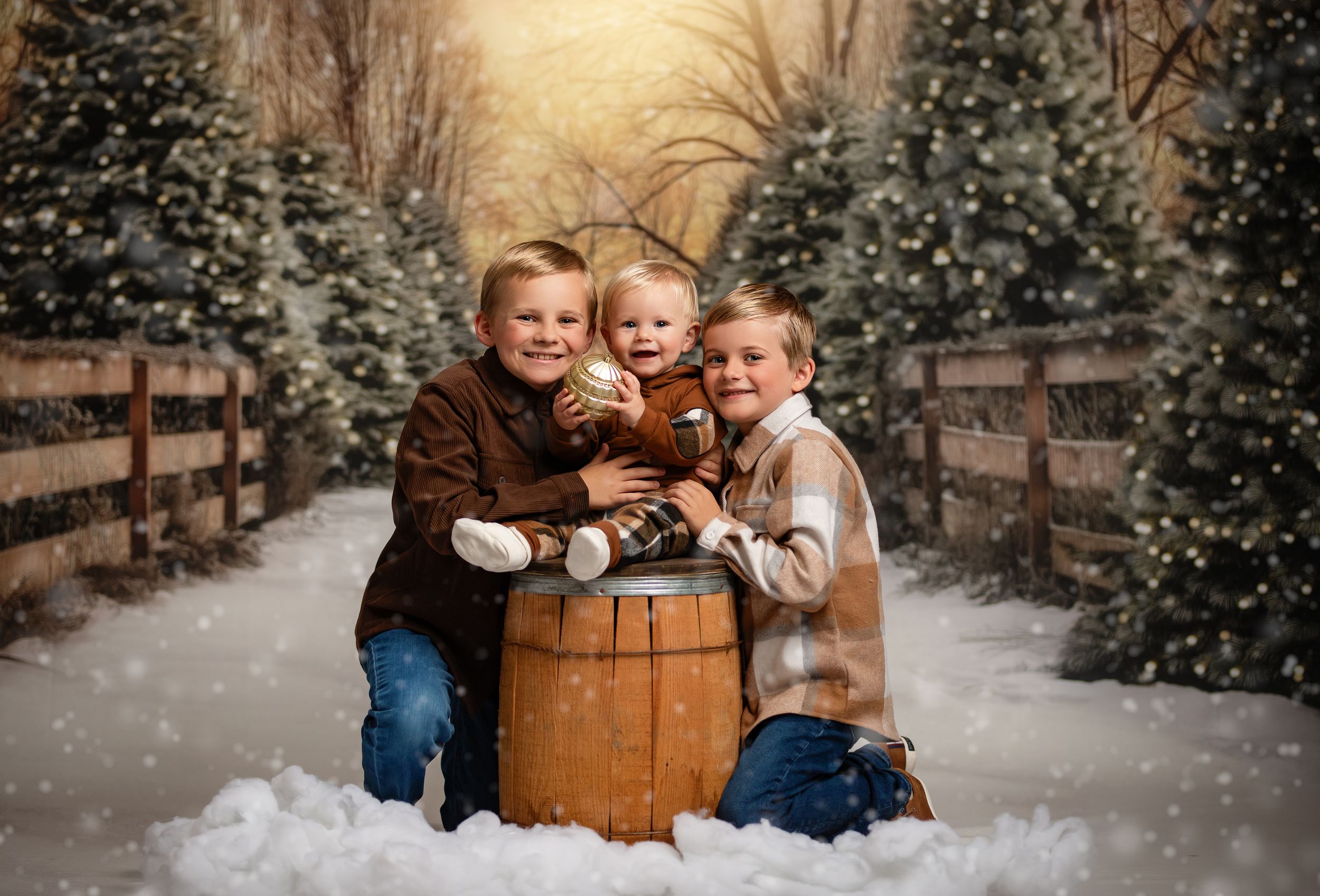 Three children in winter coats pose together around a wooden barrel in a snowy scene with evergreen trees and wooden fence.