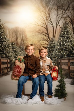 Two children sitting on a bench with Christmas lanterns in a snowy winter scene with pine trees and a wooden fence backdrop.