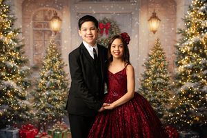 Elegant couple in formal attire poses in front of decorated Christmas trees and stone wall with twinkling lights.