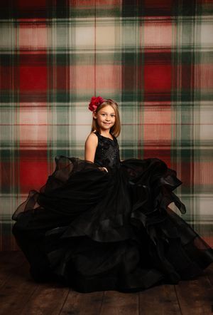 A young child poses in an elegant black ballgown against a red and green plaid backdrop.