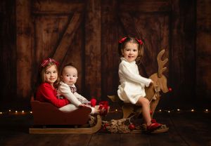 Two children in red holiday outfits sit together on a rustic wooden sleigh in a barn setting with twinkle lights.