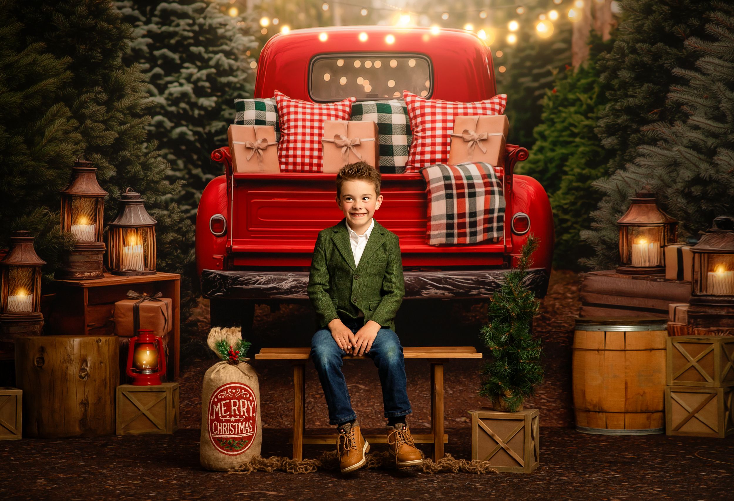 Boy in green blazer sits before red truck with Christmas gifts, lanterns, and pine trees in festive holiday photo.