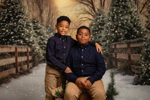 Two young boys in matching navy shirts and khaki pants pose together on a wooden bridge surrounded by snowy evergreens.