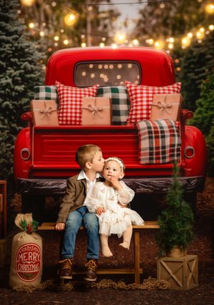 Two children sit on a bench before a vintage red truck with Christmas gifts, surrounded by festive lights and evergreen trees.