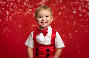 A small child in a red holiday outfit and white shirt poses against a festive red background with sparkling lights.