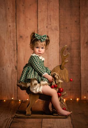 A child in a green plaid dress sits on a rustic wooden bench with Christmas lights and reindeer decor in the background.