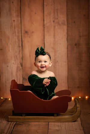 Small child wearing green velvet holiday outfit sits in decorative wooden sleigh with twinkling lights in background.