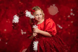 A young child in a sparkly red dress poses with white pom poms against a festive red backdrop with bokeh lights.