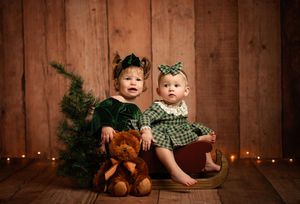Holiday portrait with small children in green outfits sitting with teddy bear and Christmas tree against wooden backdrop.