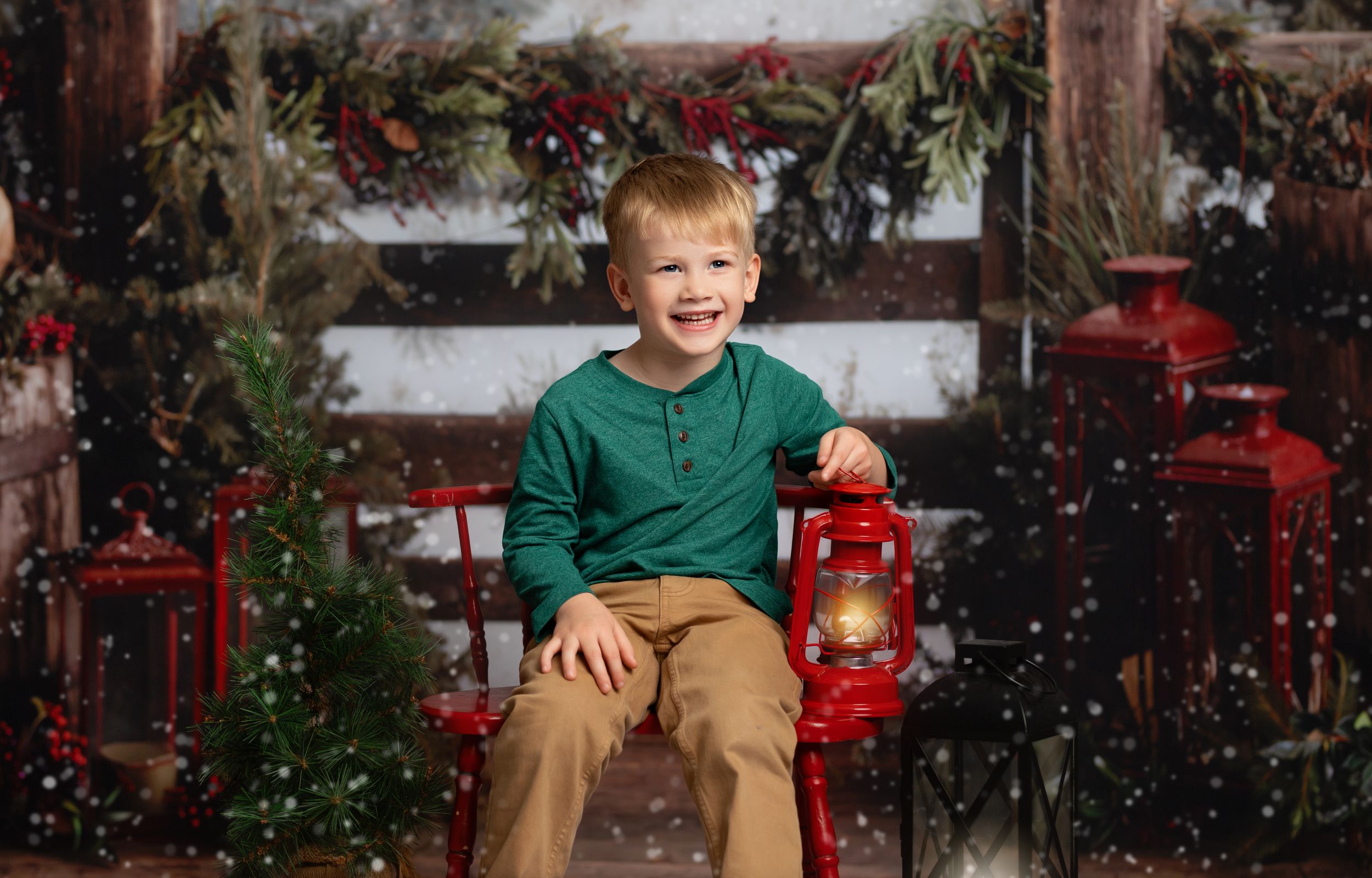 A smiling boy in a green shirt holds a red lantern on a festive Christmas-themed bench with holiday decor.