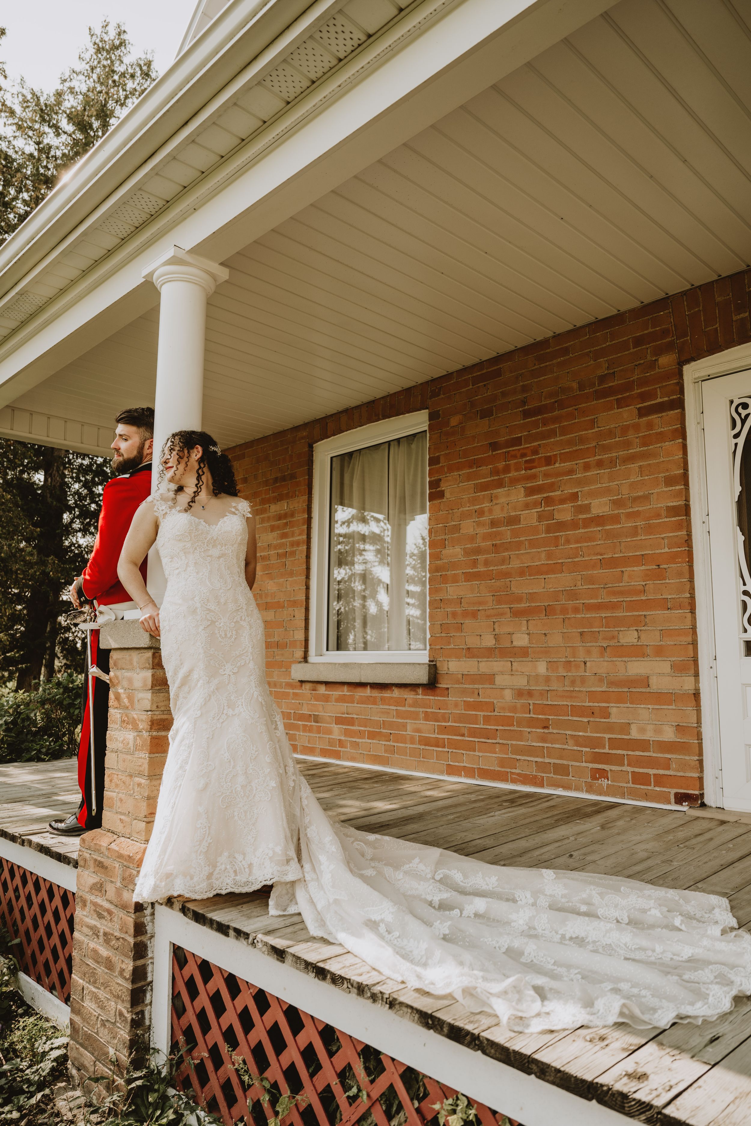 Bride in lace gown stands on porch of brick house with trailing dress.