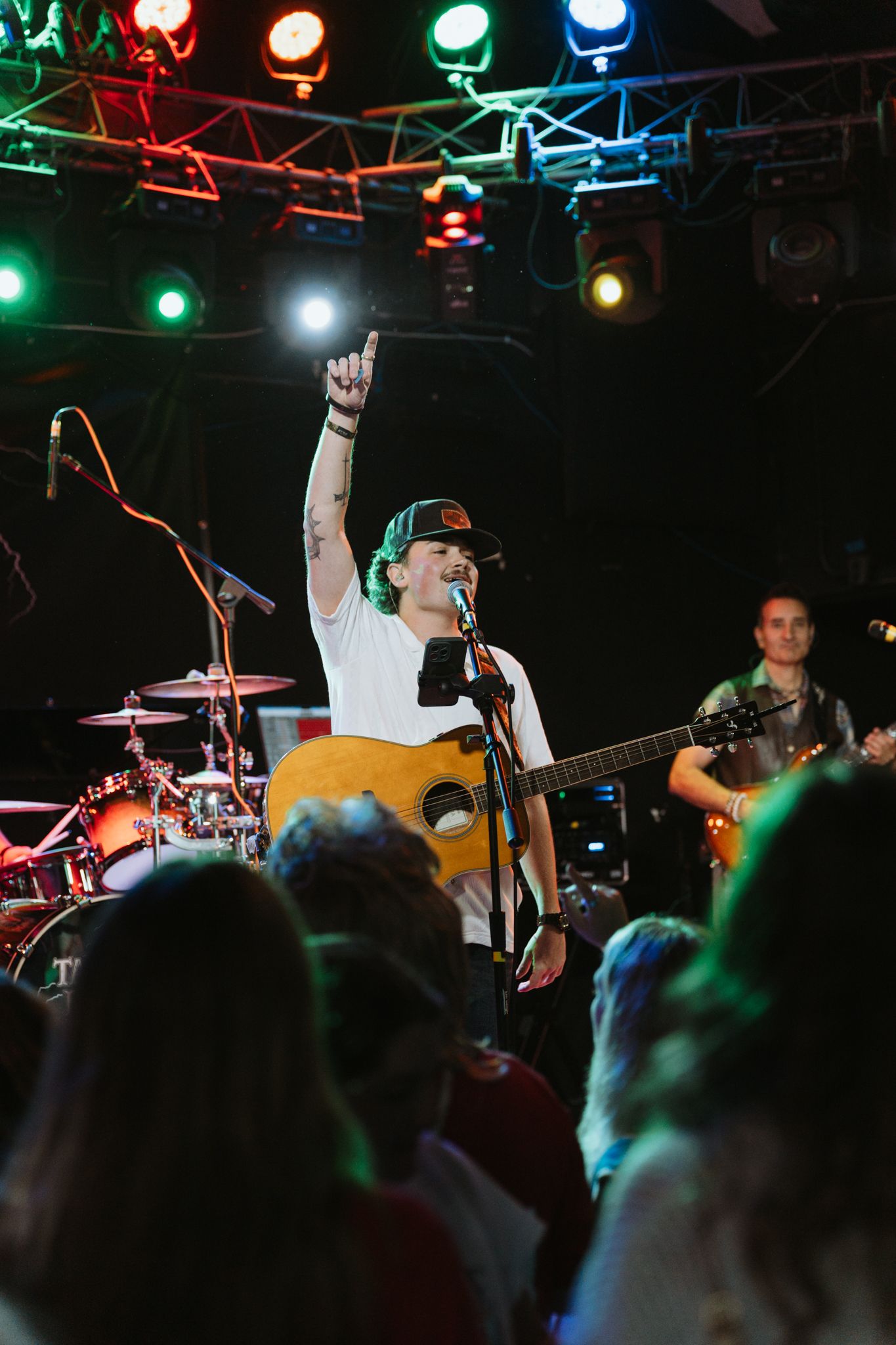 Musician plays acoustic guitar on stage with colorful lights, raising one hand to crowd during live concert performance.