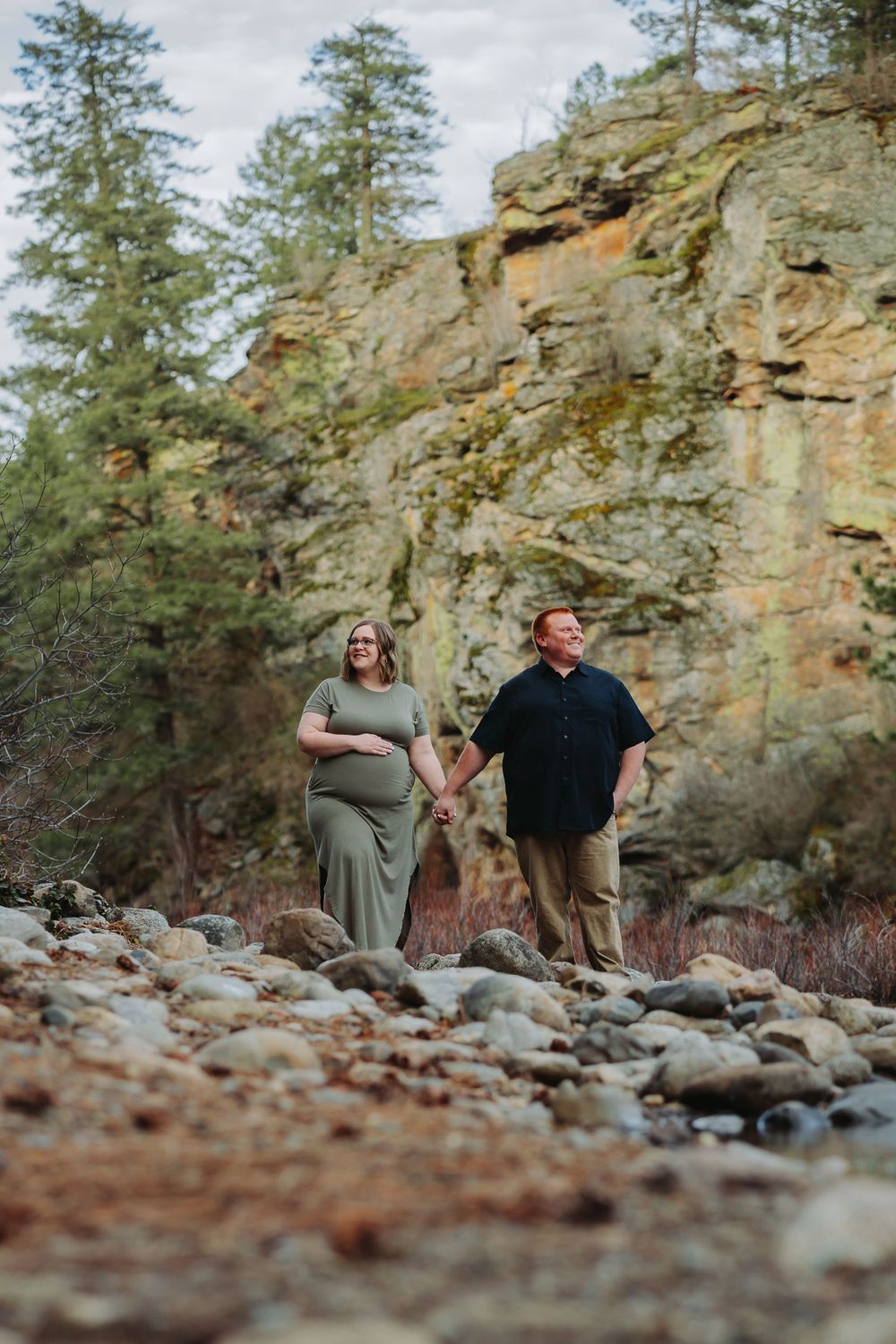 A pregnant couple walks on a rocky path in the woods, enjoying their post falls maternity session together.