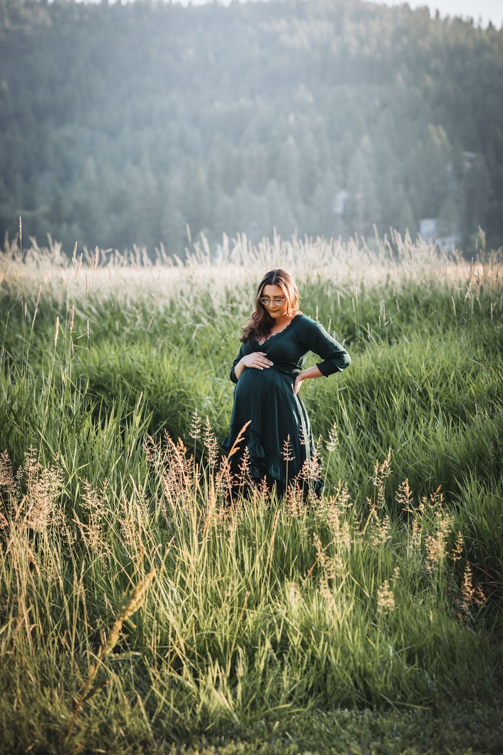 A pregnant woman in a green dress stands gracefully in a field within the Saltese Uplands, surrounded by nature's beauty.